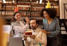 Homem assoprando as velas de um bolo de aniversário, enquanto duas colegas de trabalho, usando chapéus de festa, o surpreendem e batem palmas.