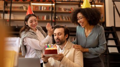 Homem assoprando as velas de um bolo de aniversário, enquanto duas colegas de trabalho, usando chapéus de festa, o surpreendem e batem palmas.