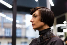 Mulher com corte de cabelo curto em estilo moderno, sendo atendida em salão de beleza, com máscara e capa protetora.