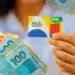 Imagem ilustrativa do Bolsa Família mostrando uma mão segurando o cartão do programa junto a notas de reais de diferentes valores, com destaque para as cédulas de 100, 50 e 20 reais.