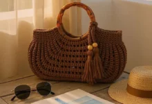 Bolsa de crochê com alça de bambu sobre mesa de madeira, com óculos de sol, chapéu e caderno.