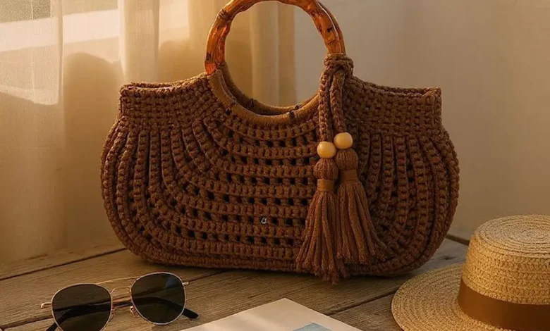 Bolsa de crochê com alça de bambu sobre mesa de madeira, com óculos de sol, chapéu e caderno.