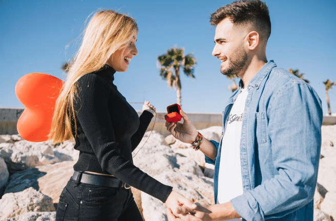 Homem fazendo pedido de casamento para mulher segurando balão vermelho em formato de coração ao ar livre