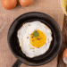 Ovo frito com gema mole na frigideira, rodeado por ovos crus, cascas, gemas separadas e utensílios. Ilustra a preparação de ovos fritos e o segredo do vinagre.