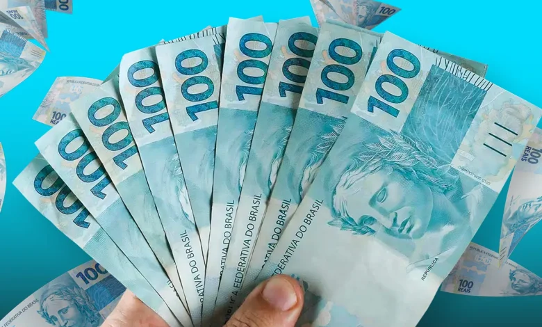 Mão segurando um leque de cédulas de 100 reais, representando o saque de até R$ 6.220 na Caixa Econômica Federal com prazo final em 13 de outubro para todos os finais de CPF.