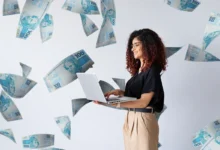 Mulher sorridente usa notebook enquanto notas de 100 reais flutuam ao seu redor, representando o recebimento indevido do auxílio emergencial.