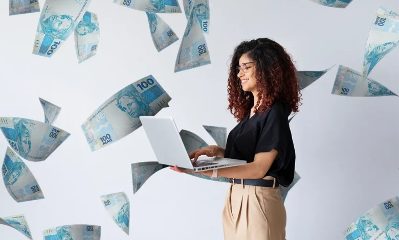Mulher sorridente usa notebook enquanto notas de 100 reais flutuam ao seu redor, representando o recebimento indevido do auxílio emergencial.