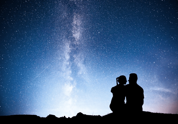 Silhueta de casal sentado sob céu estrelado com a Via Láctea visível