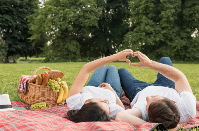 Casal deitado no chão no parque formando um coração com as mãos durante piquenique