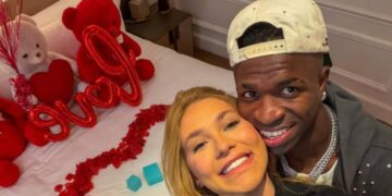 Homem e mulher sorrindo próximos à cama com decoração de ursinhos e balões vermelhos formando a palavra love