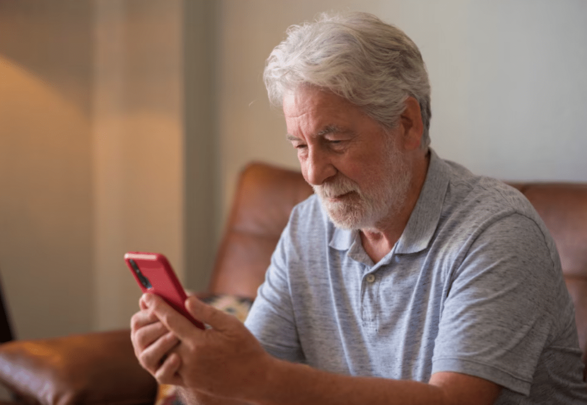 Homem idoso de camisa cinza usando celular vermelho enquanto está sentado no sofá de couro marrom
