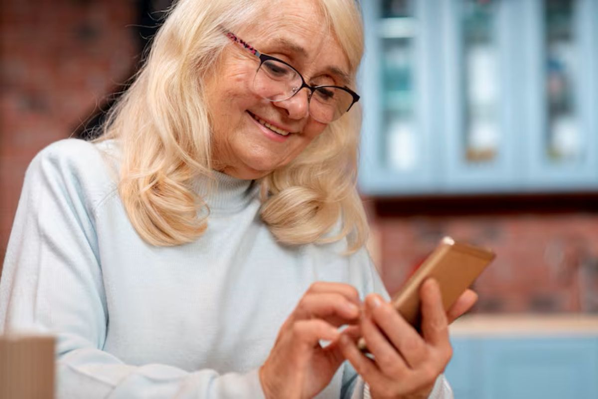 Mulher idosa de óculos e cabelo loiro sorrindo, concentrada ao interagir com um smartphone, ilustrando o envio de mensagens motivacionais.