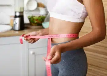 Mulher medindo a cintura com uma fita métrica, estilo saudável, associada a bebida que promete emagrecimento.