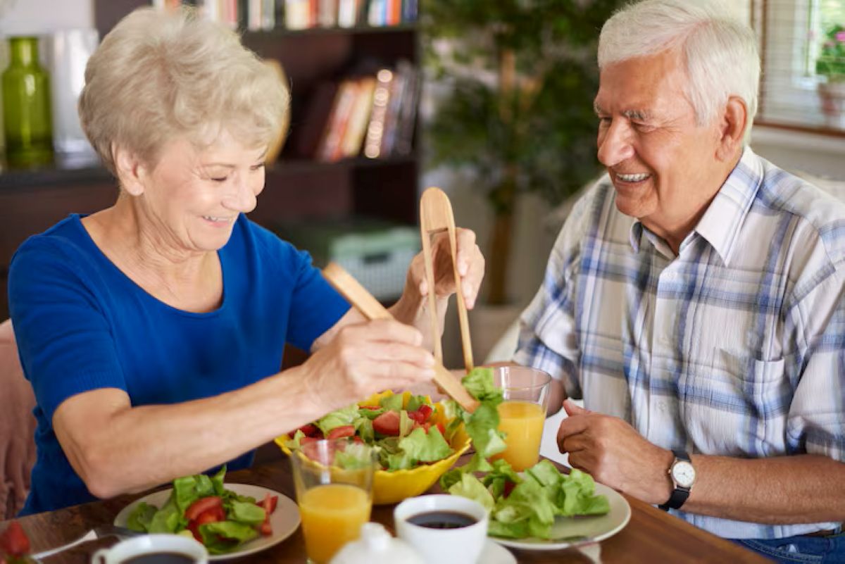 Casal idoso sorrindo em uma refeição saudável, servindo uma tigela de salada. Ilustra a importância da dieta para a saúde gástrica.