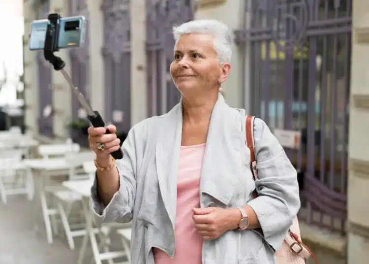 Mulher mais velha sorrindo enquanto usa um selfie stick para tirar uma foto em um ambiente urbano.