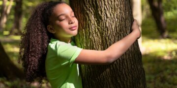 Menina de cabelos cacheados em uma camisa verde, abraçando o tronco áspero de uma árvore em uma floresta, com os olhos fechados, expressando calma e a sensação de alívio do estresse na natureza.