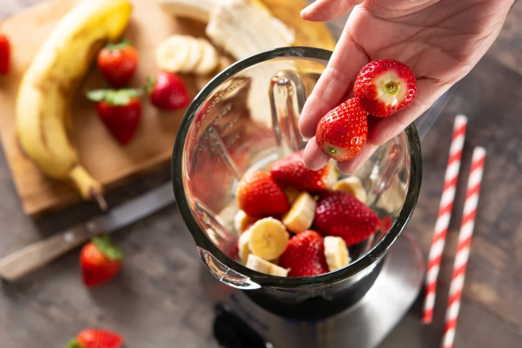 Mão colocando morangos frescos em um liquidificador com bananas fatiadas e morangos. 