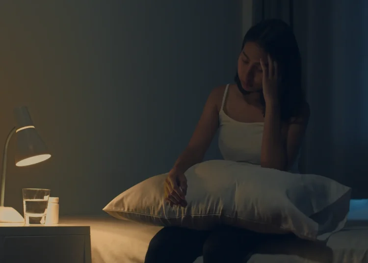 Mulher sentada na cama à noite segurando um travesseiro, com dificuldade para dormir.