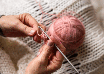 Mãos fazendo crochê com linha rosa e agulha de crochê sobre manta de lã
