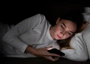 Mulher deitada na cama olhando para o celular no escuro.