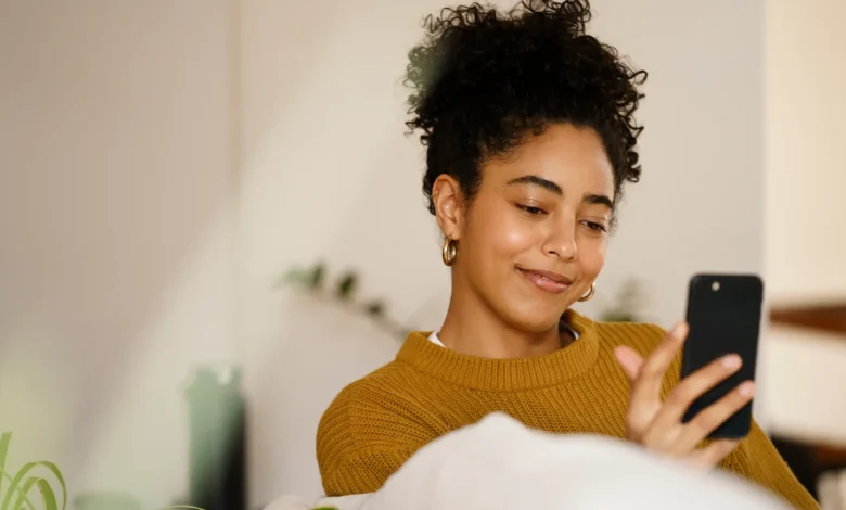 Mulher sorrindo enquanto usa smartphone, desejando boas vibrações para outubro aos amigos no WhatsApp.