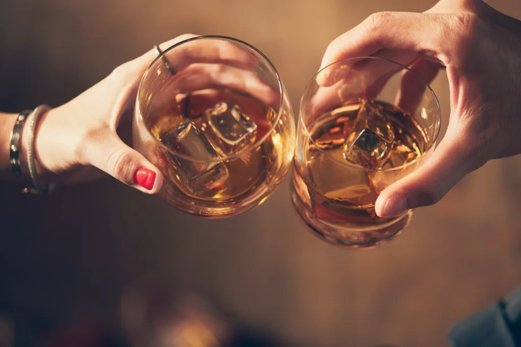 Dois copos de whisky brindando, com gelo visível, ilustrando o consumo de álcool e o perigo da ingestão incorreta de substâncias como metanol.