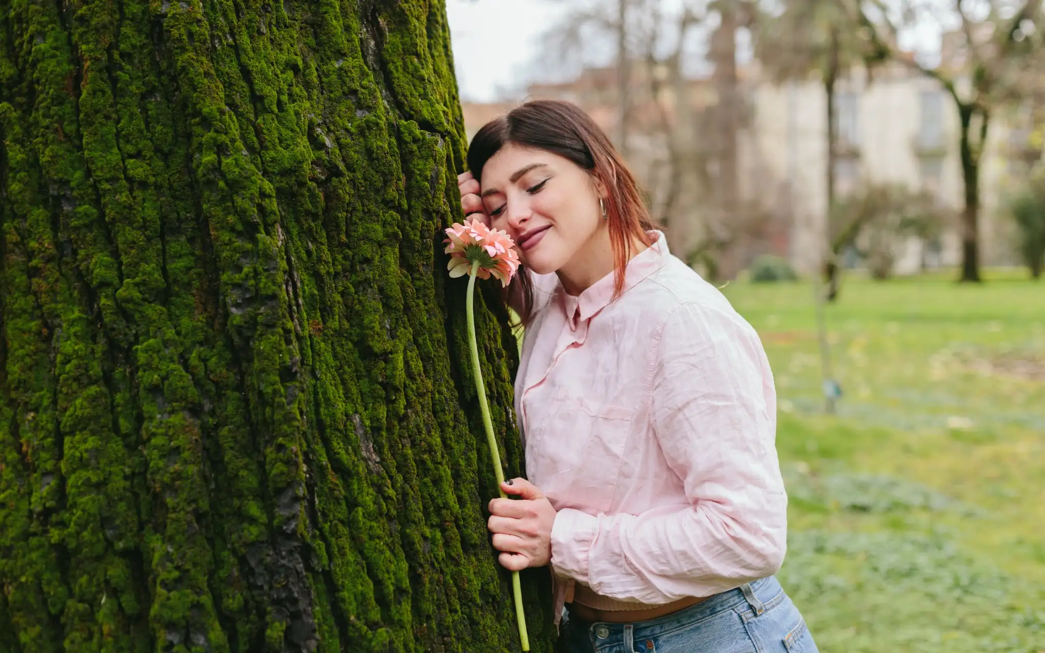 Mulher em ambiente ao ar livre, sorrindo enquanto segura uma flor próxima a uma árvore coberta de musgo.