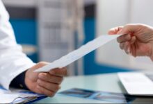 Mãos de um profissional de saúde (jaleco branco) entregando uma receita médica ou documento a um paciente. Foco na transferência da prescrição.