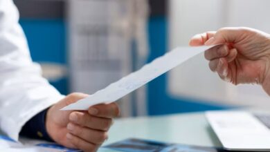 Mãos de um profissional de saúde (jaleco branco) entregando uma receita médica ou documento a um paciente. Foco na transferência da prescrição.