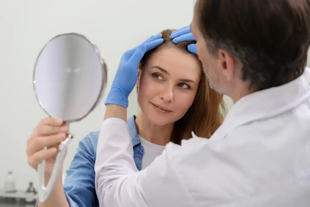 Paciente mulher sendo examinada por dermatologista sobre queda de cabelo, com a ajuda de um espelho para observar o couro cabeludo.