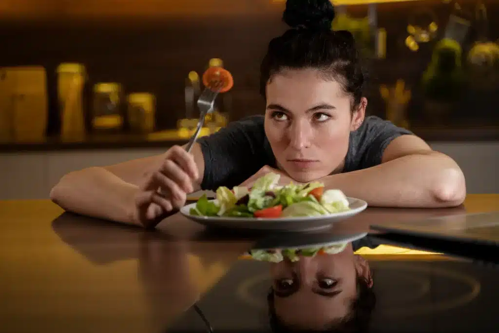 Mulher olhando com desânimo para um prato de salada, representando restrições alimentares extremas e dietas da moda.