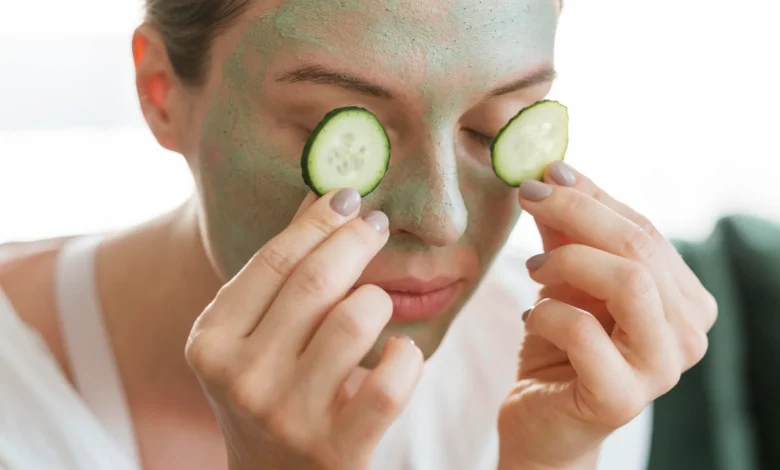 Mulher aplicando pepino sobre os olhos enquanto usa máscara facial de argila, ilustrando cuidados com a pele.