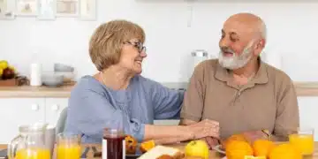 Casal sorridente tomando café da manhã saudável com frutas, suco de laranja e pães sobre a mesa.