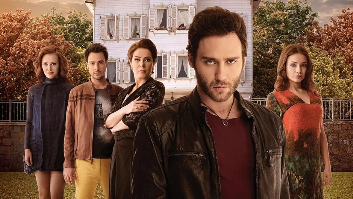 Pôster da série Em Uma História de Amor, mostrando Korkut Ali e outros personagens principais em um cenário dramático, com fundo de uma casa e árvores no outono