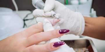 Manicure utilizando lixa elétrica para remover o alongamento de unha em gel, com profissional e cliente usando luvas de proteção em um salão de beleza.
