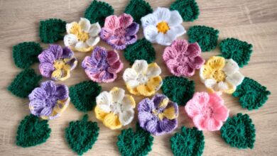 Arranjo de flores 3D de crochê e folhas em tons pastel (roxo, rosa, amarelo e branco) sobre superfície de madeira clara, ilustrando a aplicação em artesanato de moda.