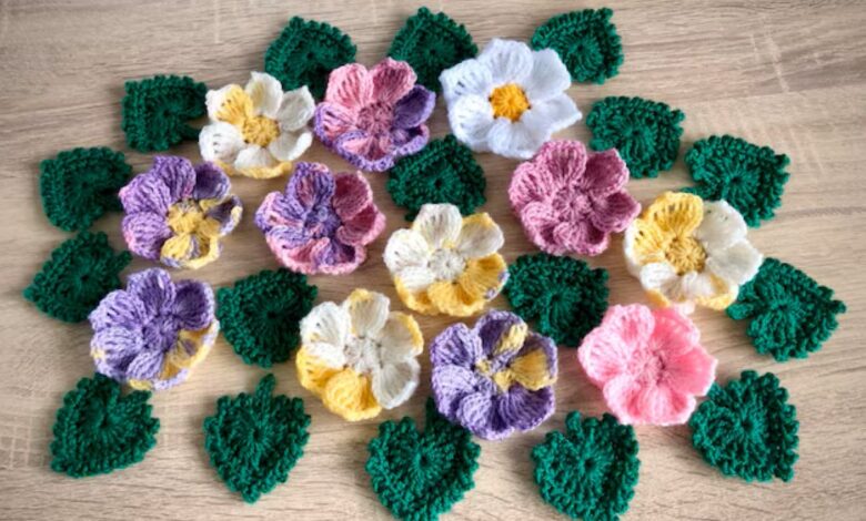 Arranjo de flores 3D de crochê e folhas em tons pastel (roxo, rosa, amarelo e branco) sobre superfície de madeira clara, ilustrando a aplicação em artesanato de moda.