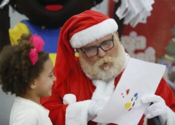 Campanha Papai Noel dos Correios 2025: Papai Noel de óculos lendo atentamente a cartinha de Natal de uma criança, simbolizando a adoção de cartas e a realização de sonhos.