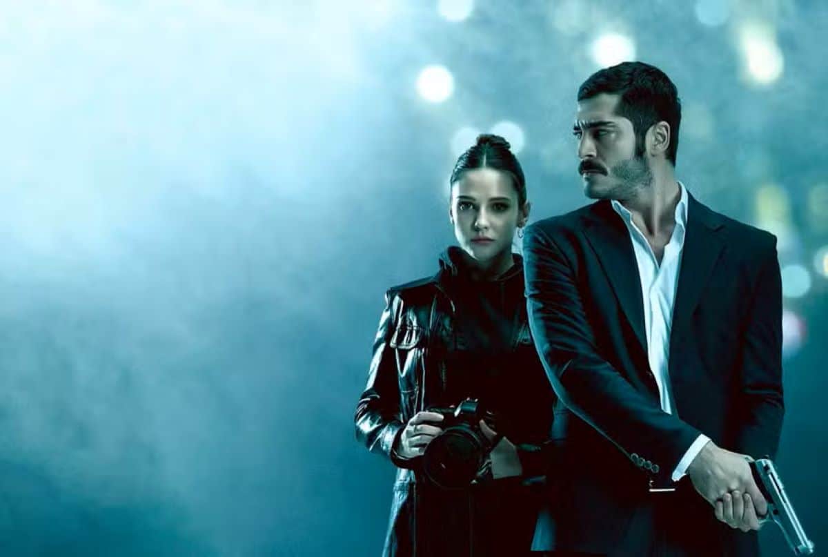 Um homem com bigode e terno, segurando uma arma, ao lado de uma mulher com câmera e jaqueta de couro, ambos com expressões sérias, sobre um fundo esfumaçado, representando a ação da novela turca 'Maraslı: O Protetor'.