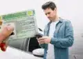 Homem ao lado de um carro consulta o celular enquanto uma Carteira Nacional de Habilitação brasileira é exibida em primeiro plano.