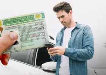 Homem ao lado de um carro consulta o celular enquanto uma Carteira Nacional de Habilitação brasileira é exibida em primeiro plano.