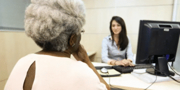 Aposentada idosa conversando com atendente em uma mesa de escritório, em frente a um computador, representando atendimento bancário ou financeiro.