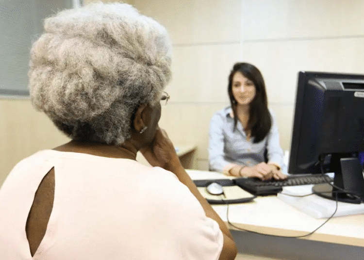 Aposentada idosa conversando com atendente em uma mesa de escritório, em frente a um computador, representando atendimento bancário ou financeiro.