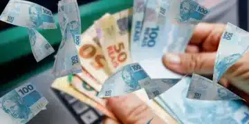 Notas de 100 reais e 50 reais flutuando ao redor de uma mão segurando dinheiro, com um caixa eletrônico ao fundo. Relacionado ao auxílio de R$ 250 aprovado no RS.