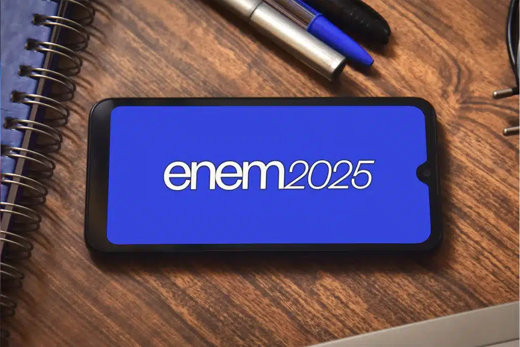 Imagem do ENEM 2025 em um smartphone sobre uma mesa de estudos, com canetas e cadernos ao fundo.