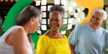 Três idosos sorridentes conversando, mulher de amarelo sorrindo no centro