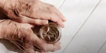 Mãos de idoso segurando pote transparente cheio de moedas de euro sobre mesa branca