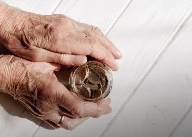 Mãos de idoso segurando pote transparente cheio de moedas de euro sobre mesa branca