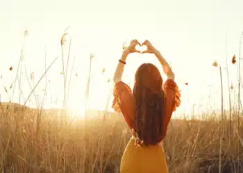 Mulher de costas no campo ao pôr do sol, fazendo um gesto de coração com as mãos. Uma representação de amor próprio e autocuidado.