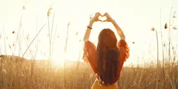 Mulher de costas no campo ao pôr do sol, fazendo um gesto de coração com as mãos. Uma representação de amor próprio e autocuidado.